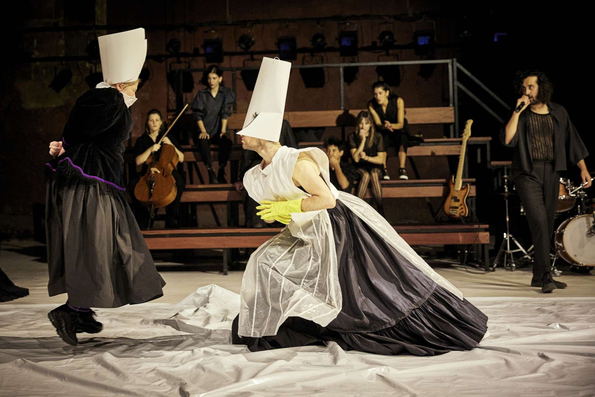 Deux personnages habillés en cardinaux évoluent sur une bâche transparente. En fon de scène, un gradin avec des musiciens et musiciennes dessus. 