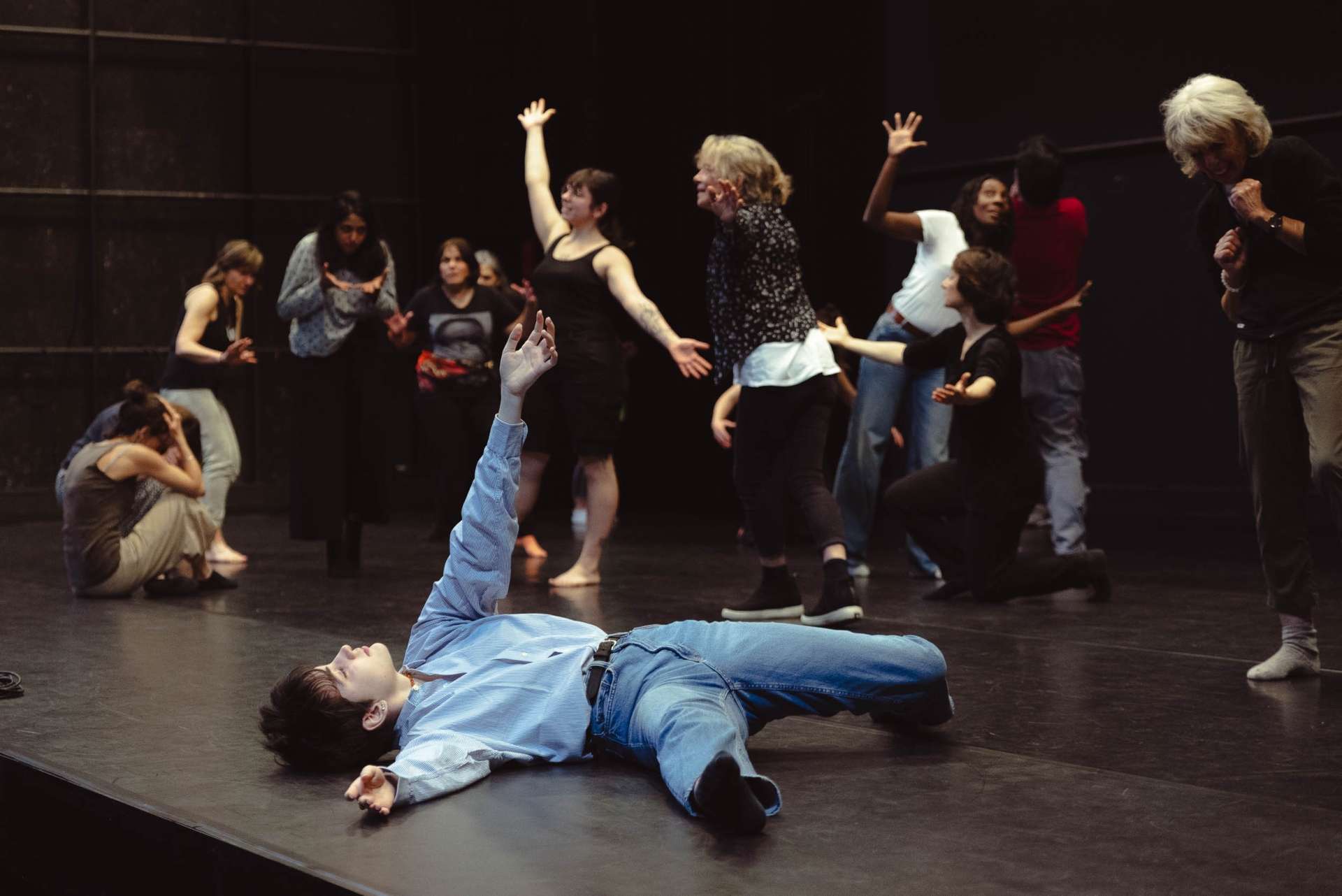 En bord de scène, une amateure est allongée avec un bras levé. En fond de scène, les autres participants au projet danse ou marche. 