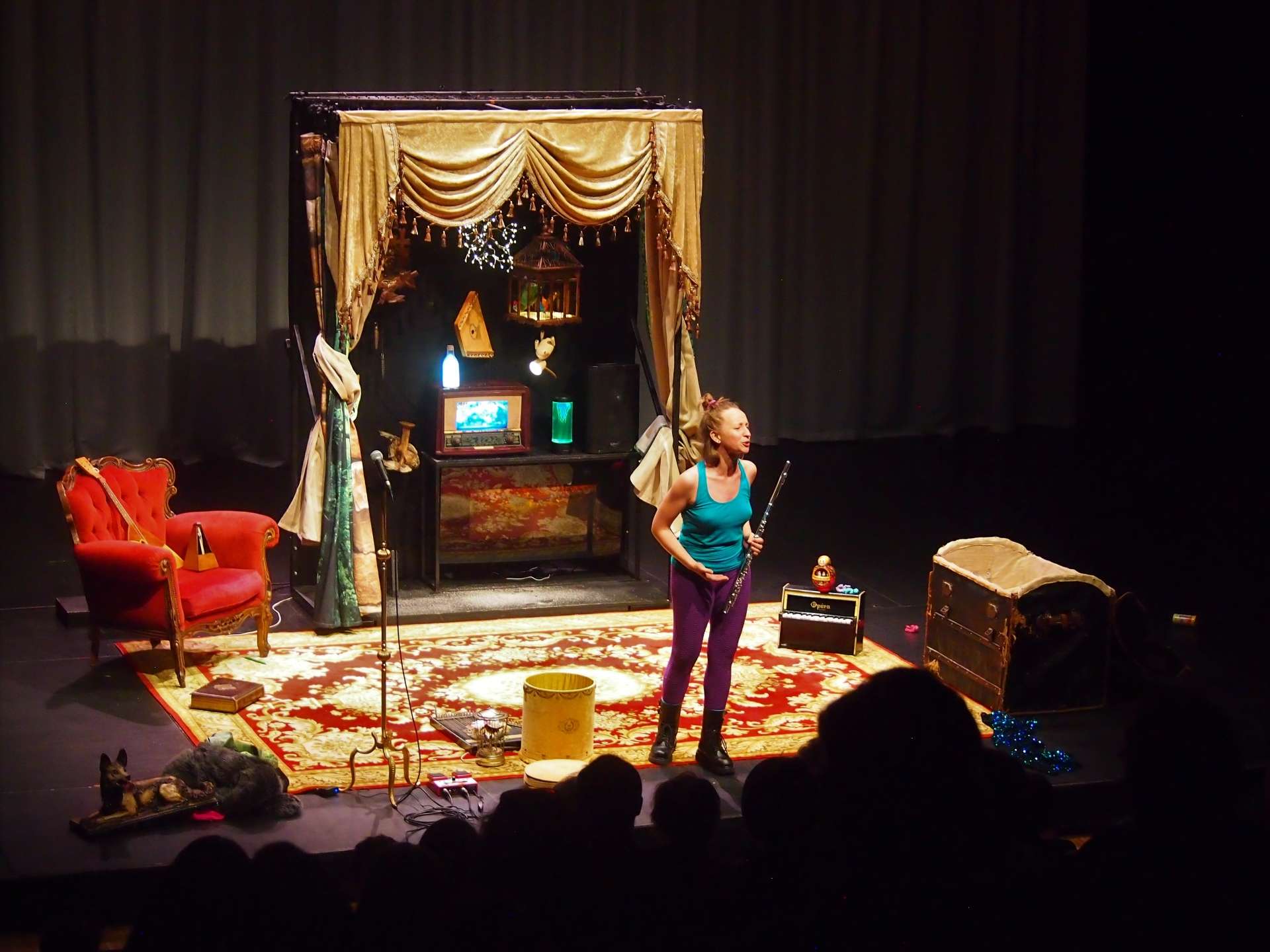 Le décor de la chambre d'Anya : fauteuil rouge, tapis persan, rideau ancien, coffre... et Anya au premier plan avec sa flûte traversière à la main. 